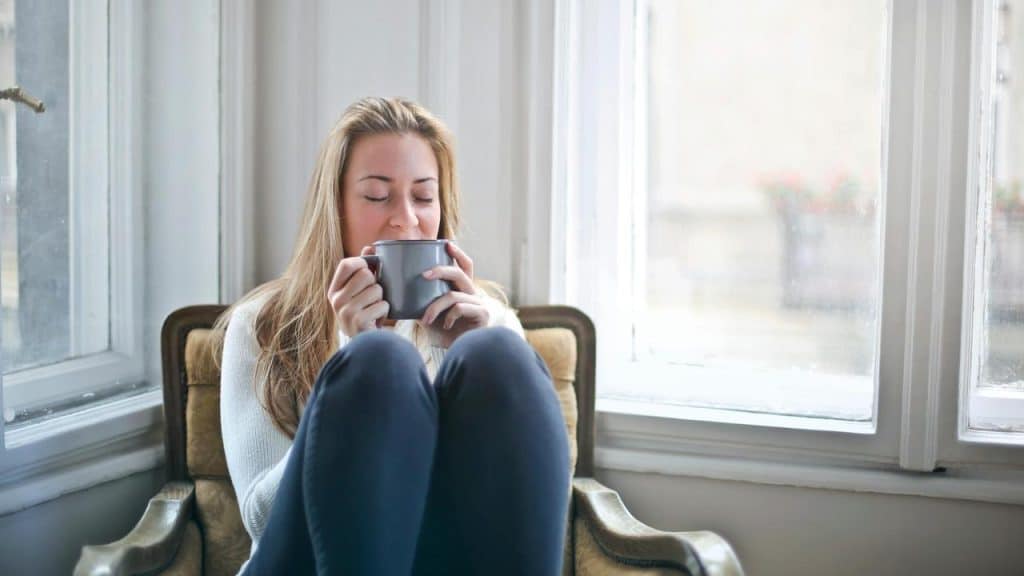 A woman sitting by a window while holding a mug with closed eyes.
