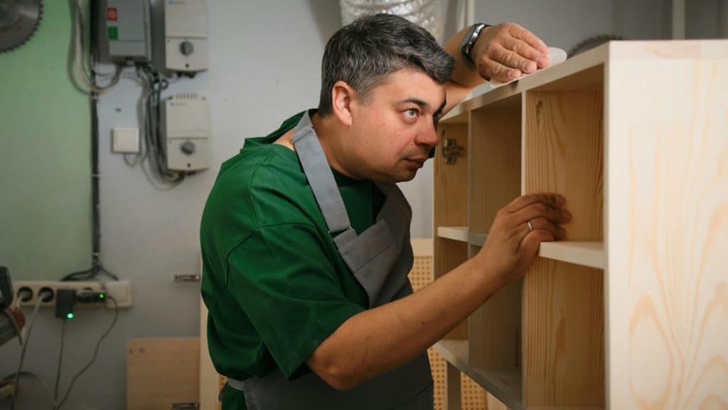 A craftsman in a green shirt and apron is carefully inspecting a wooden shelving unit in a workshop.