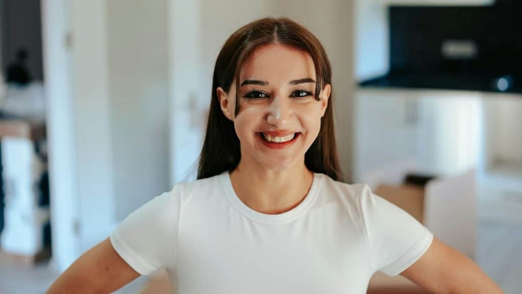 A smiling young woman wearing a white t-shirt is standing in a living room.