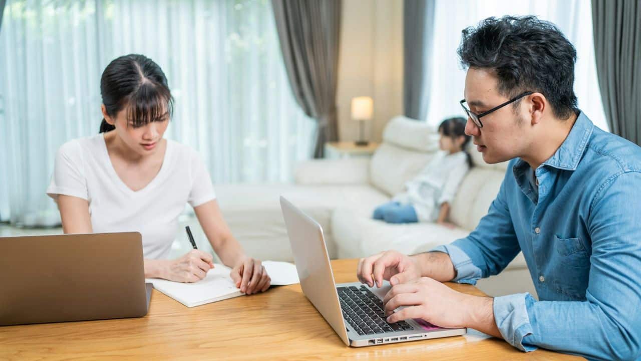 A man and woman work on a laptop and notebook at a table in a bright living room.
