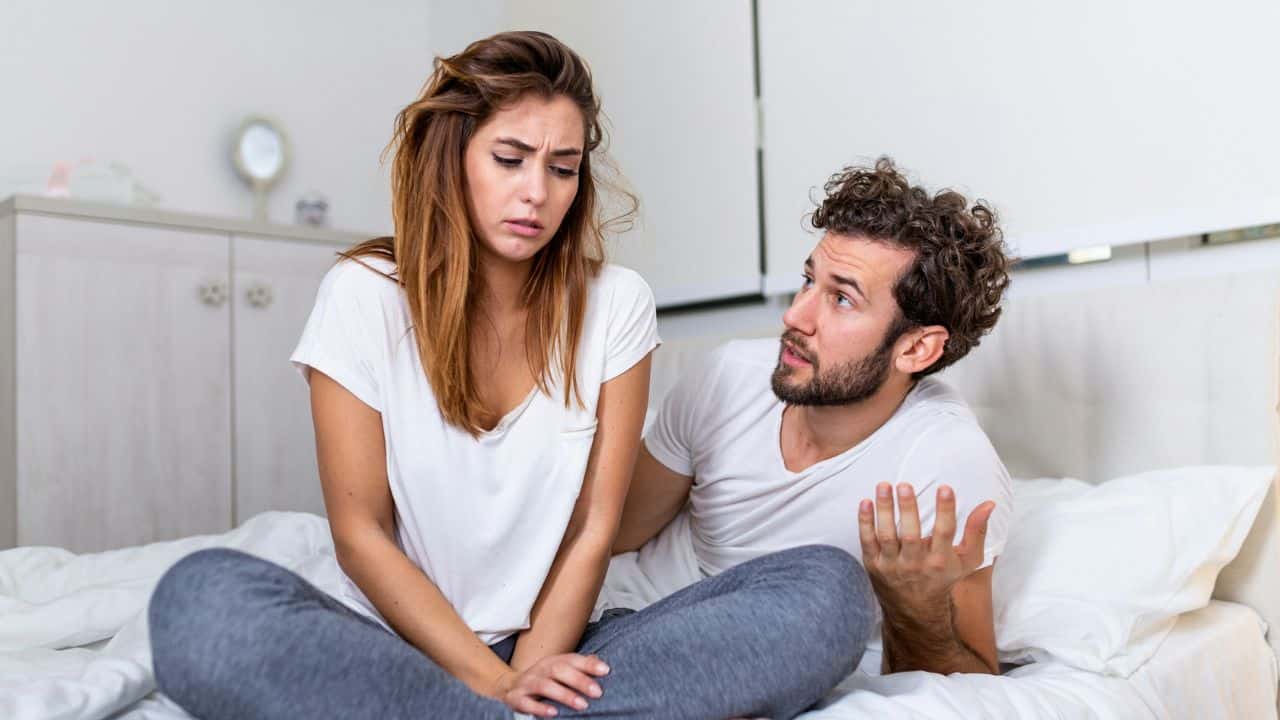 A distressed woman sits on a bed while a man talks to her with his hands raised.