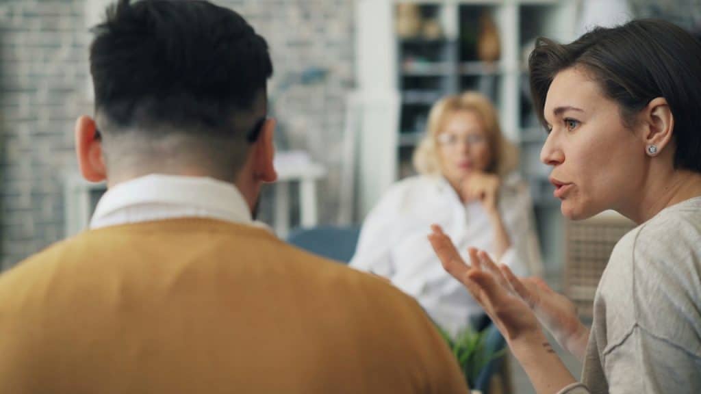 Woman arguing and gesturing across a table towards a man, with a therapist or counselor visible in the blurred background.