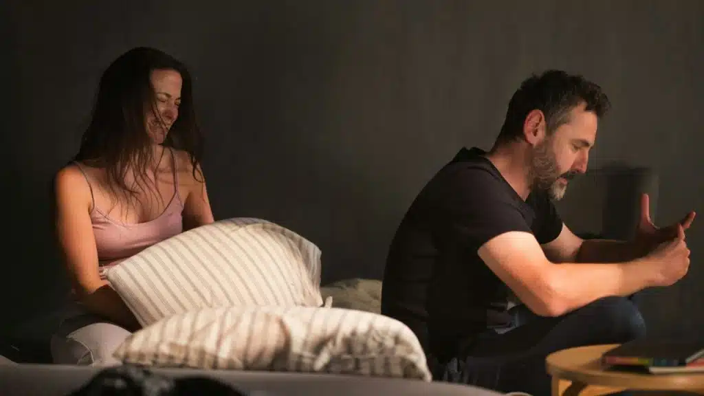 A woman smiles while holding a pillow next to a distressed man sitting beside her.