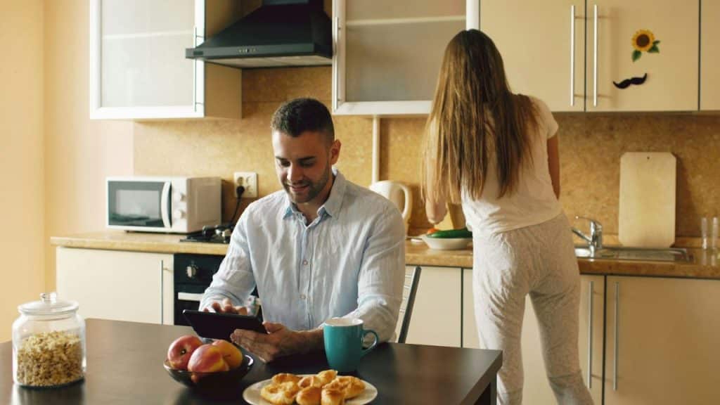 A man uses a tablet at a table while a woman works in the background kitchen.