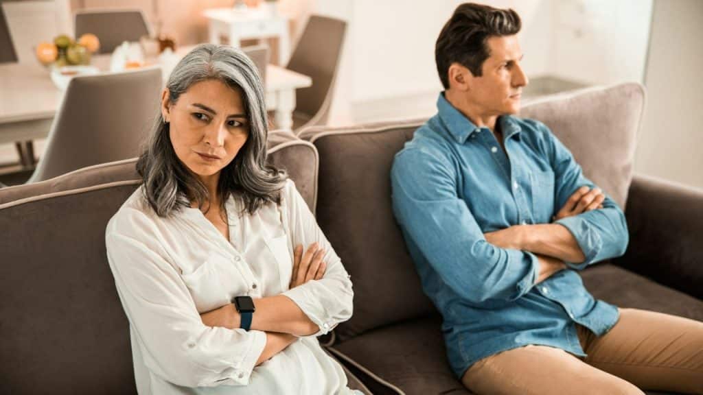 An angry-looking couple sits on a couch with their arms crossed, looking away from each other.