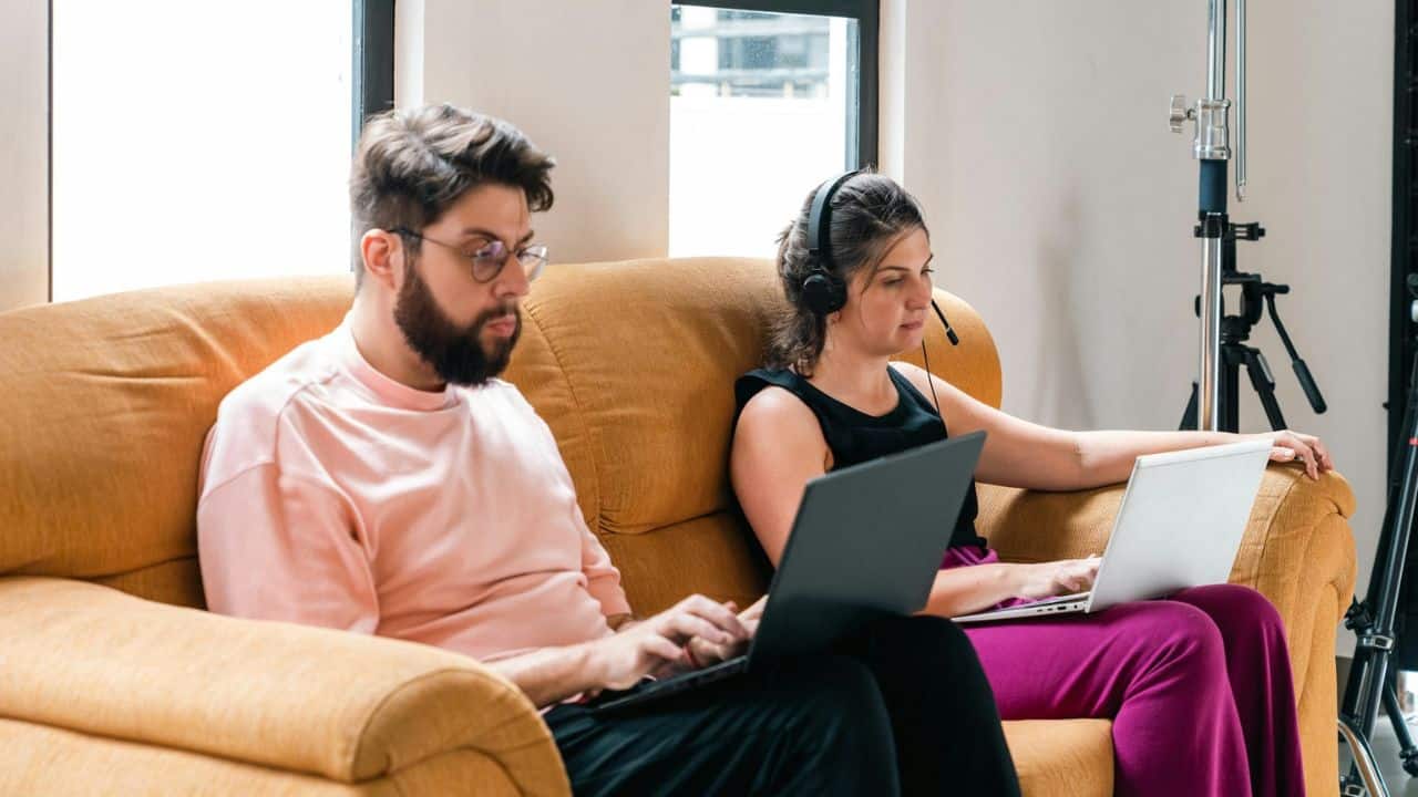 Two people sit on a couch using laptops, one wearing a headset.