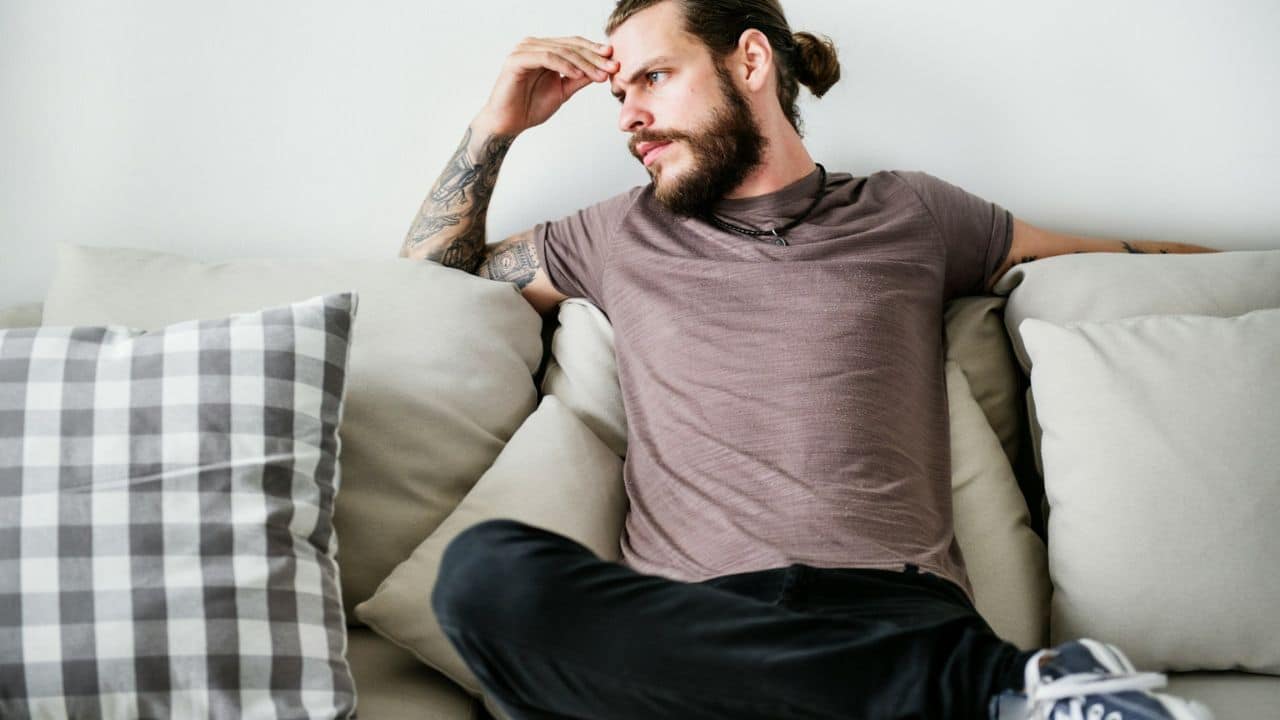 A bearded man with tattoos and a topknot sits on a couch looking away.