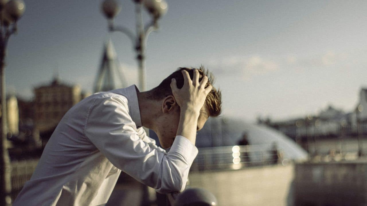 A man in a white shirt with his hands on his head leans against a railing outdoors.