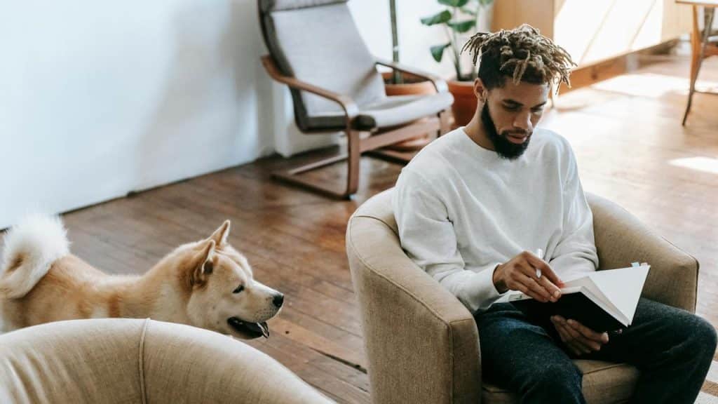 A man sitting in a chair reading a book with a dog beside him.