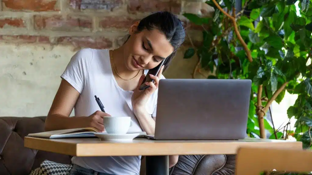 A woman talking on the phone while writing notes at a table with a laptop.