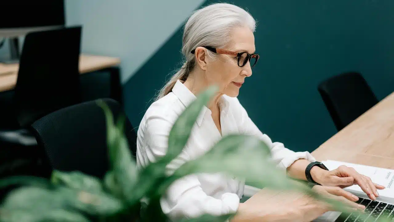 An older woman typing on a laptop in an office.