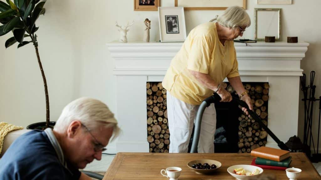 An older woman vacuums a fireplace while an older man sits at a table in the foreground.