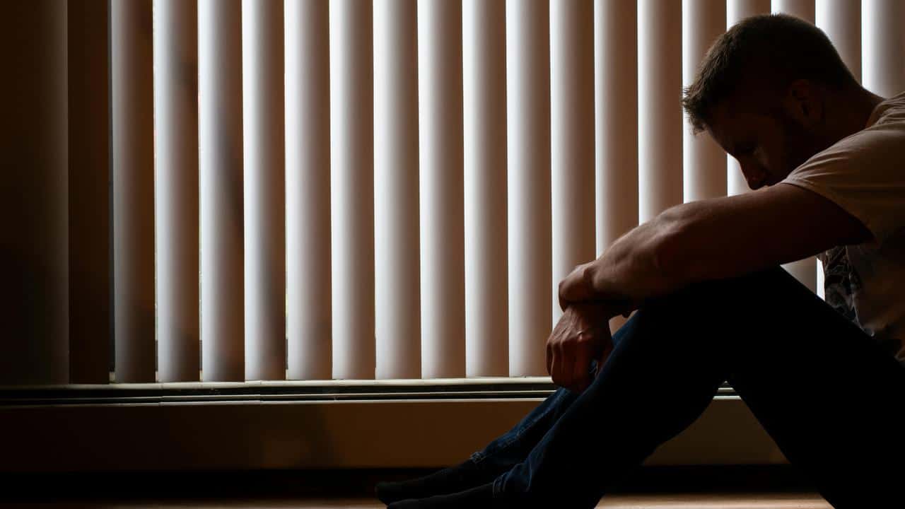 A man sitting on the floor in silhouette with his head bowed near window blinds.