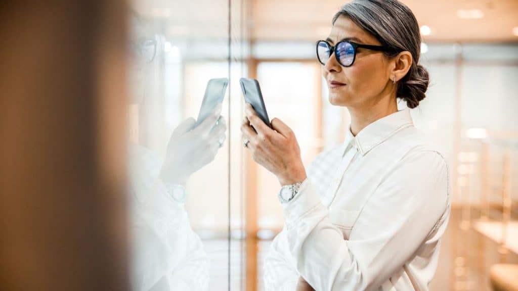 Middle-aged woman with gray hair and glasses using a smartphone indoors.