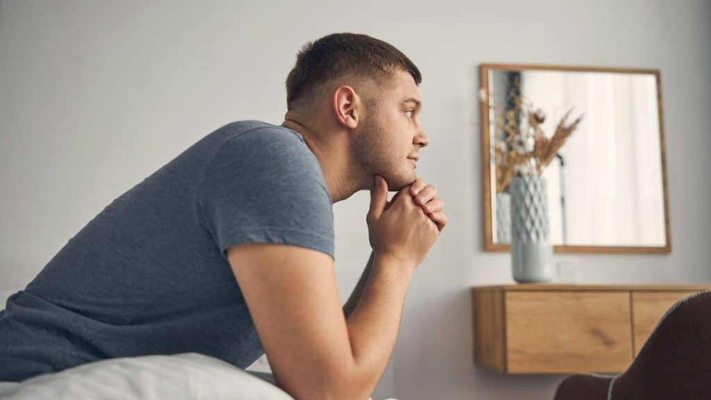 Young man with short hair sitting on a bed with hands clasped under his chin.