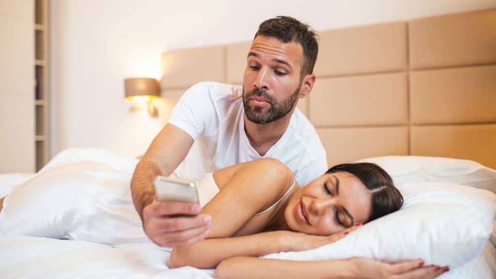 A man is leaning over a woman in bed, looking at the phone she is holding.