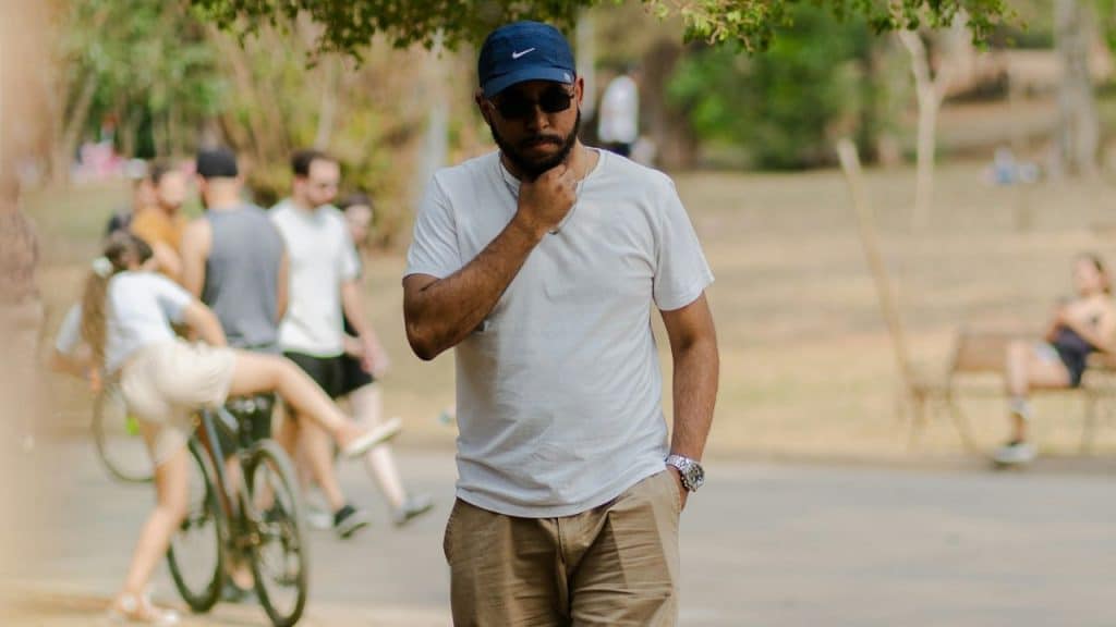 A man in sunglasses and a cap stands alone in a sunny park with people in the background.