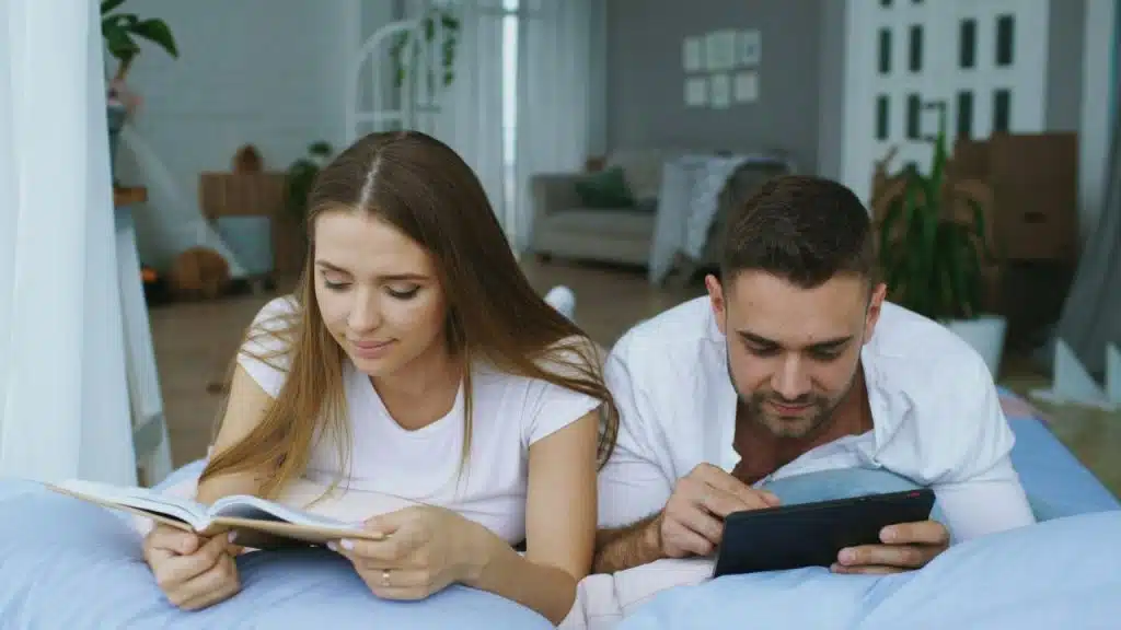 A man and woman lie on a bed; the woman reads a planner, the man uses a tablet.