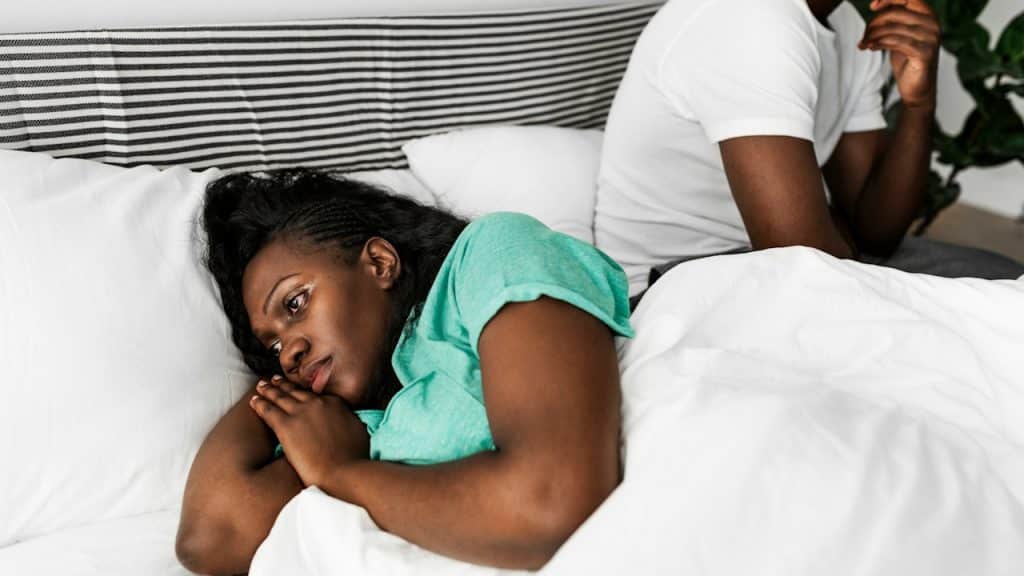 A woman lies awake in bed looking unhappy, with a man sitting behind her looking thoughtful or distant.