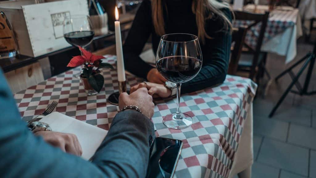 A couple is holding hands across a table set with wine glasses, a candle, and a checkered tablecloth during a romantic dinner.