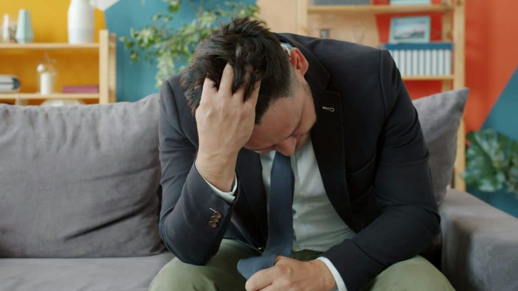 Man in a suit sitting on a couch, holding his head in distress or exhaustion.
