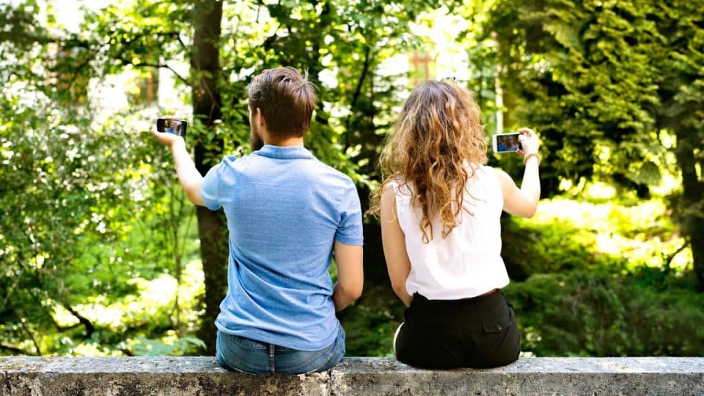 A man and woman are sitting outdoors, backs to the camera, taking photos on their phones.