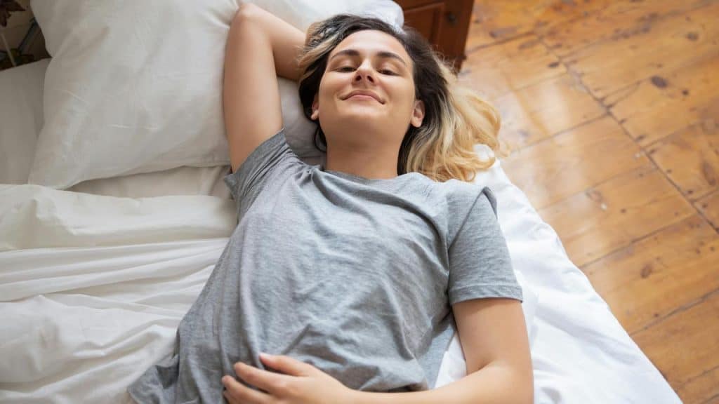 A woman in a gray shirt lies on a bed smiling contentedly.