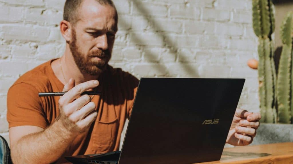A bearded man looks intently at a laptop, holding a pen.