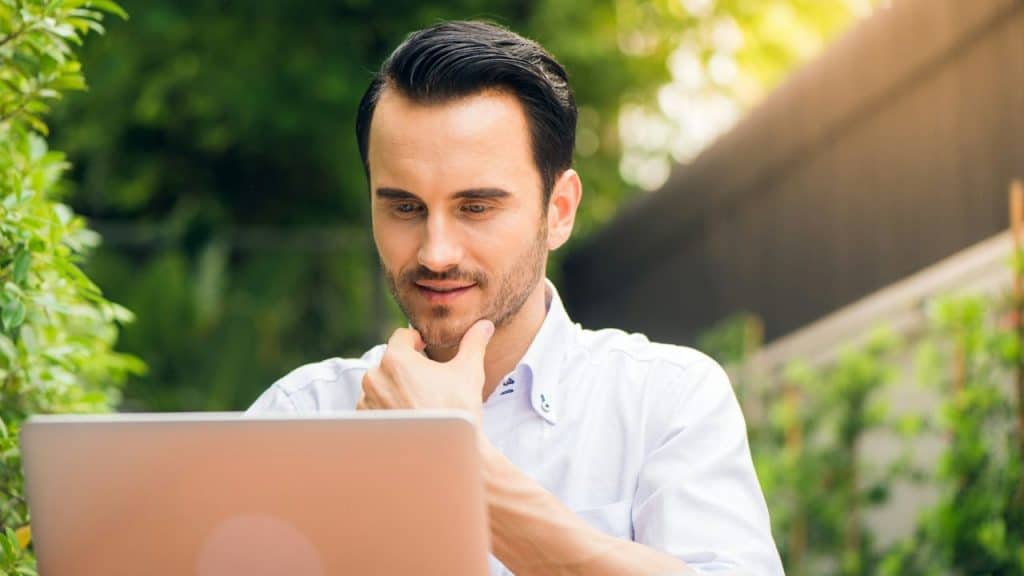 Man in a white shirt with his hand on his chin, looking at a laptop outdoors.
