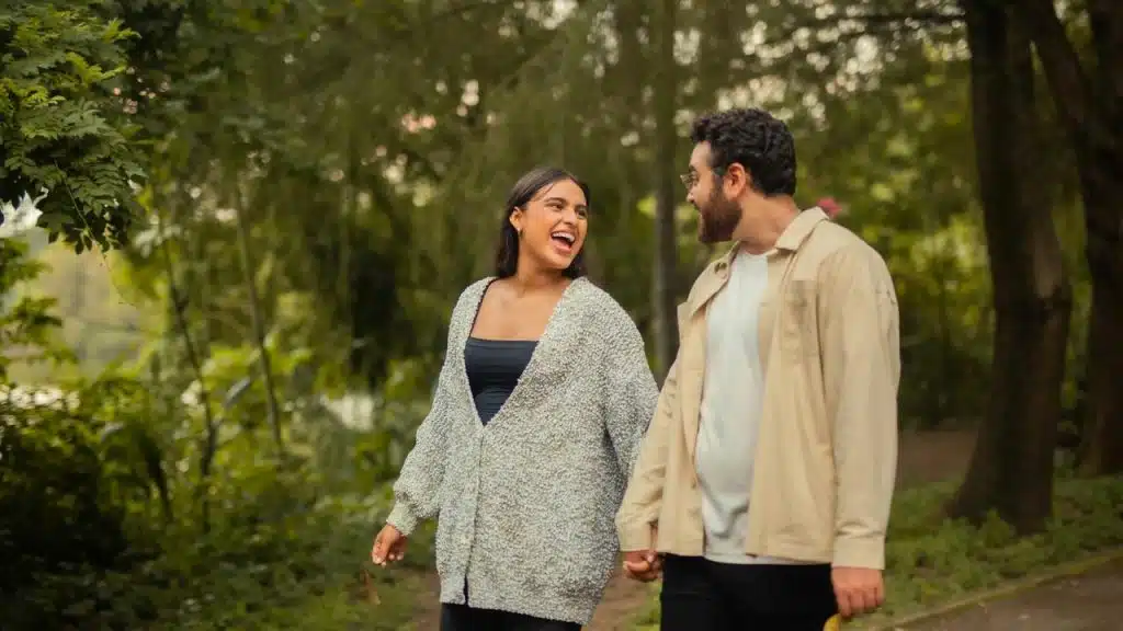 A couple walking hand in hand and smiling in a park surrounded by trees.