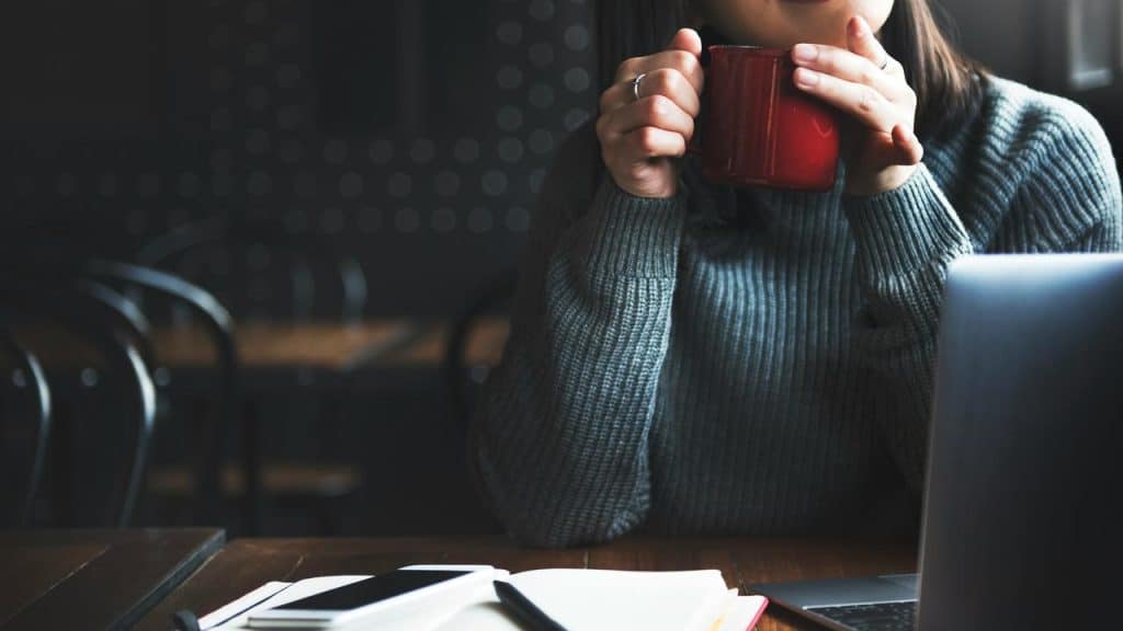 A person sitting at a table with a laptop, holding a red mug.