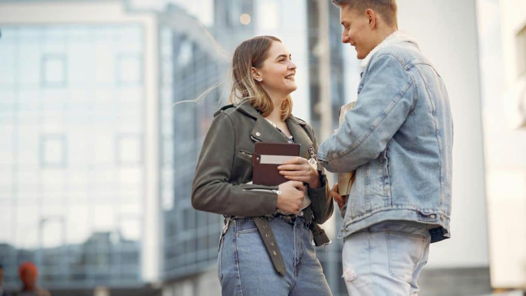 A couple smiling and talking while standing outside with books.
