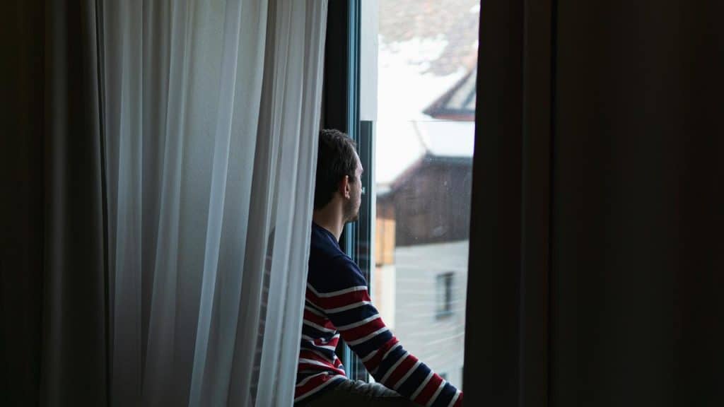 A man sitting by a window looking outside through sheer curtains.