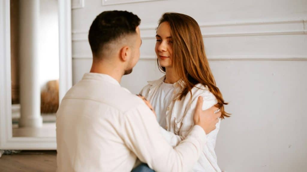 A man puts his hands on a woman's shoulders while looking at her in a white room.