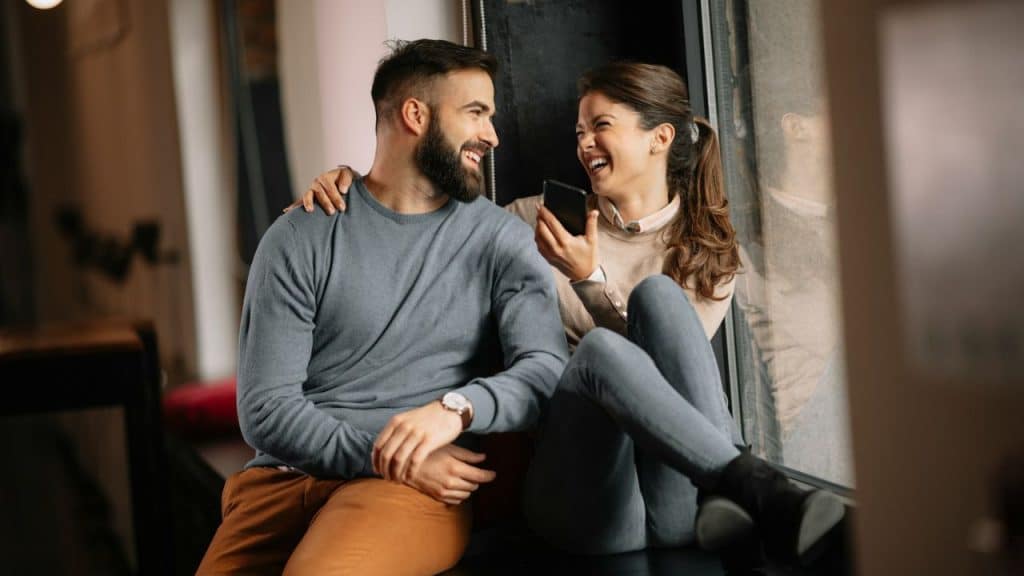 Happy bearded man and woman sitting together by a window, laughing and looking at a cell phone.