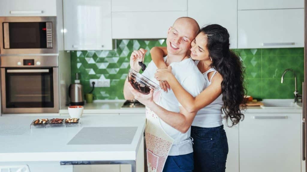 Woman with long dark hair hugging a smiling bald man holding a bowl in a kitchen.