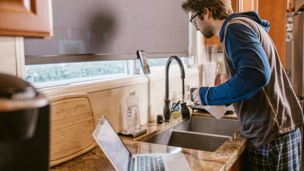 A man standing at a kitchen sink, watching something on his phone near a laptop.