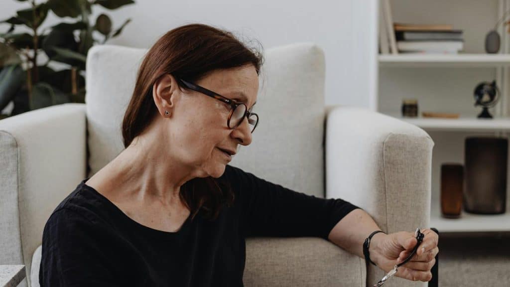 An older woman sitting on the floor beside a chair, holding her glasses thoughtfully.