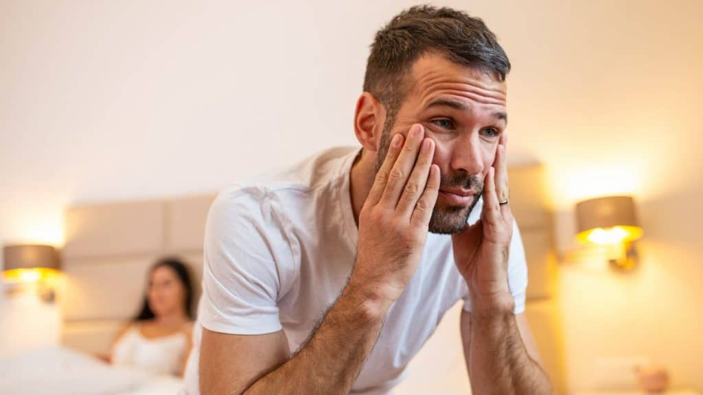 A man sits up in bed, looking troubled, holding his face with both hands, with a woman blurred in the background.