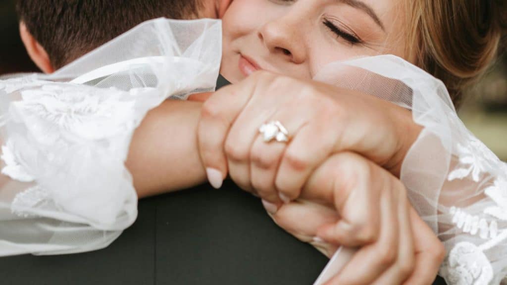 A bride with closed eyes smiles while hugging her partner tightly.