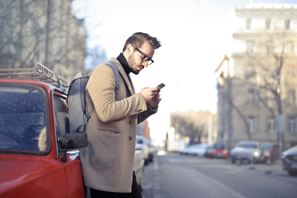 Man in Beige Coat Holding Phone Leaning on Red Vehicle