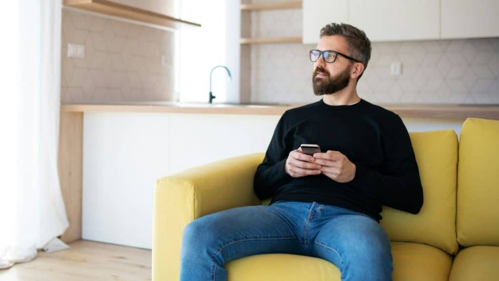 Bearded man with glasses holding a smartphone while sitting on a yellow couch.