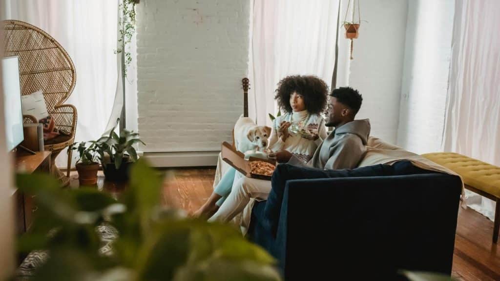 A couple sitting on a couch eating pizza with a dog beside them.