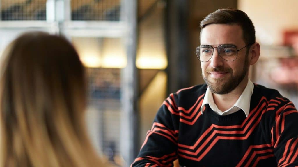 A man in a patterned sweater and glasses smiles at a blurred person.