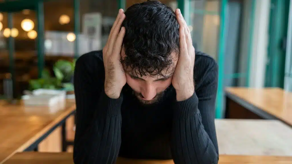 A distraught man sits at a table with his head bowed and hands on his head.