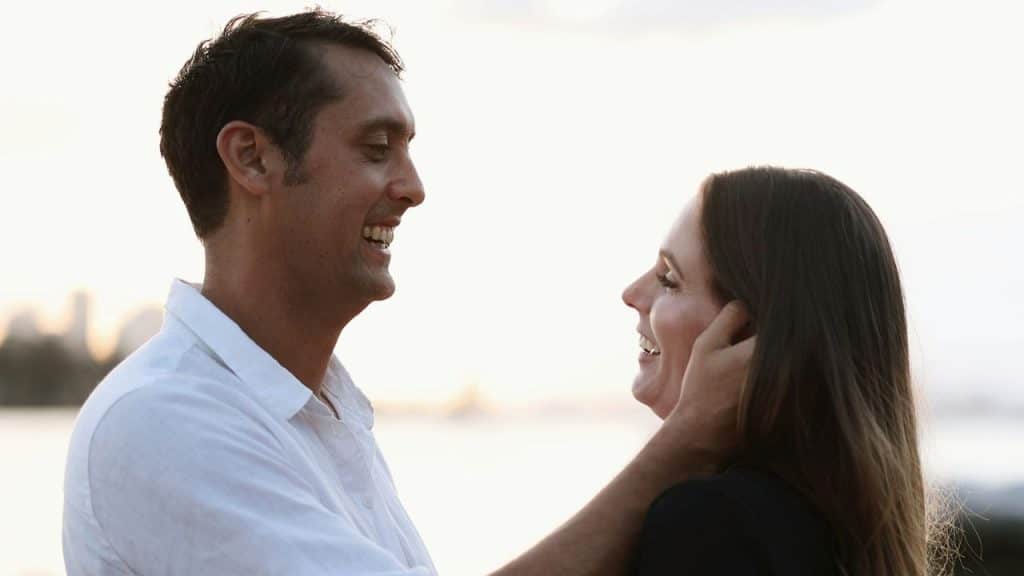 A smiling man gently cups the face of a smiling woman outdoors.