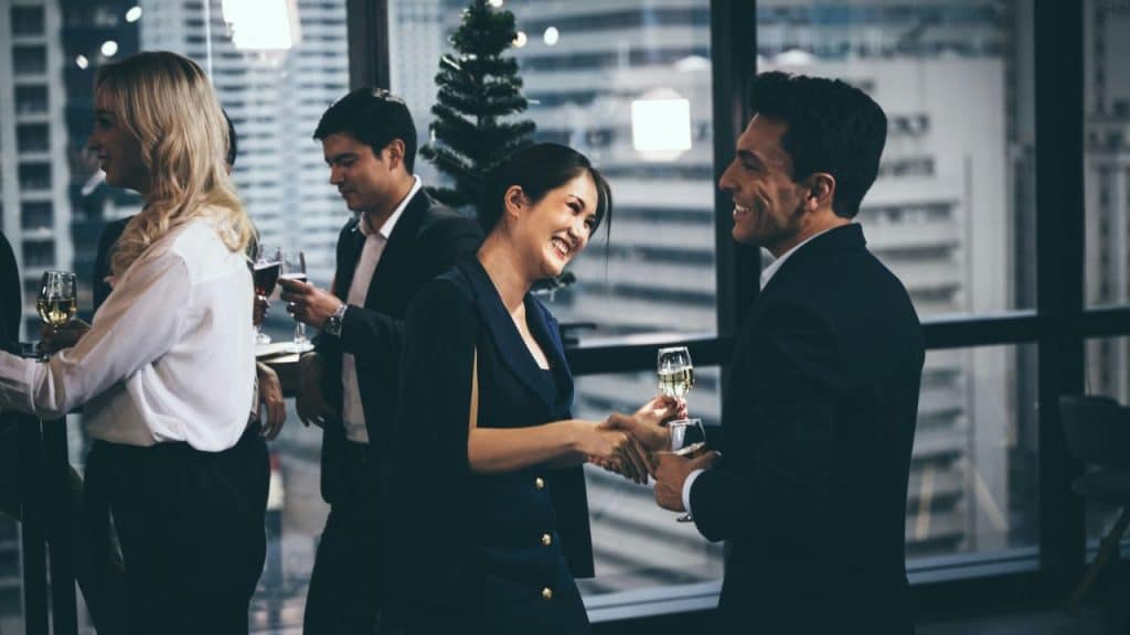 Smiling professionals in suits and business attire are shaking hands and drinking wine at a networking event in an office.