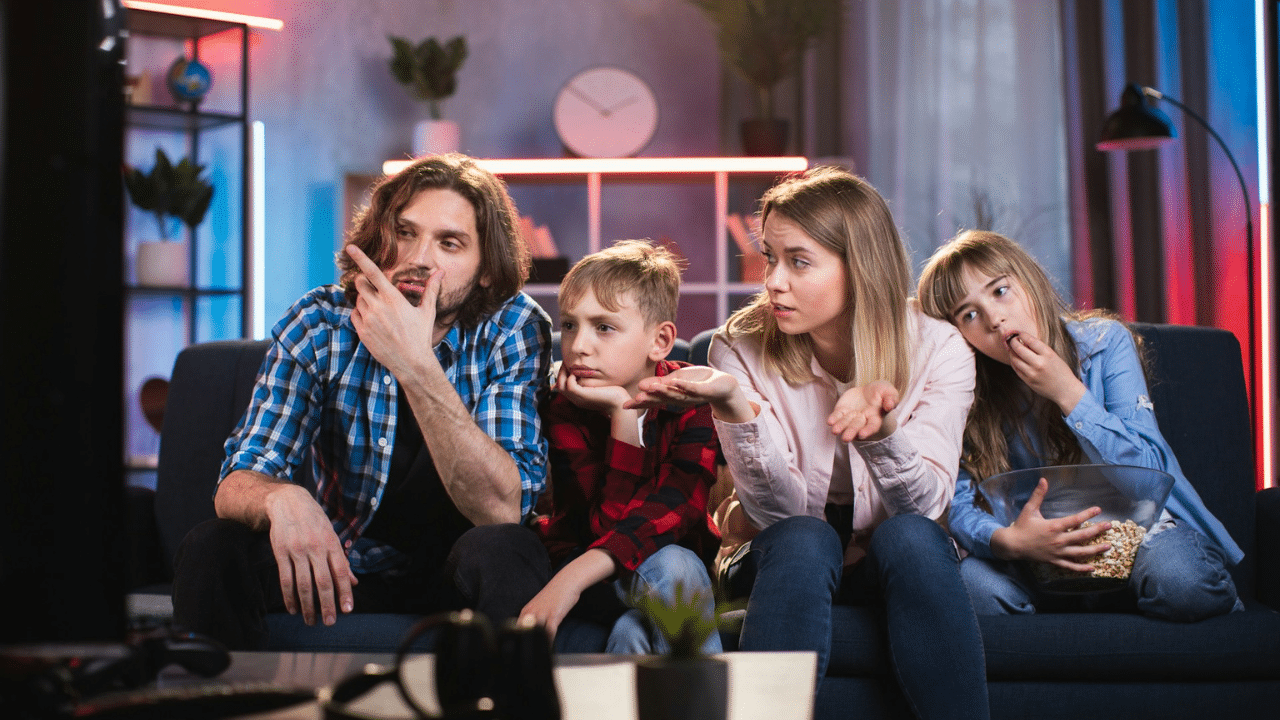 A family of four sitting on a sofa watching television with expressions of boredom or disagreement.