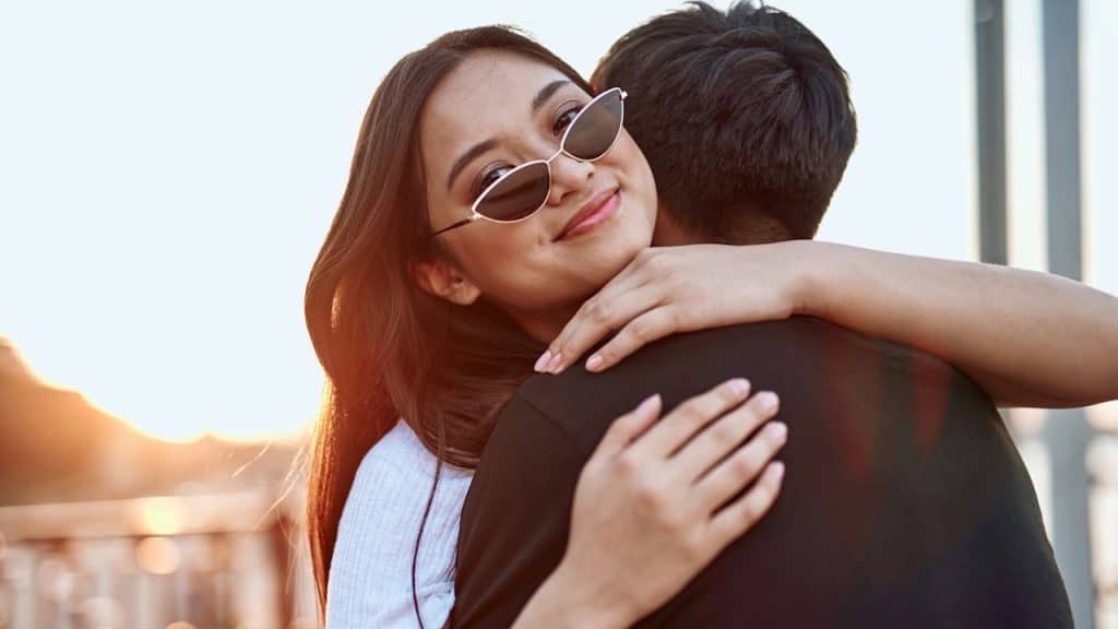 A smiling young woman wearing sunglasses is embracing a man and looking back at the camera during sunset or golden hour.