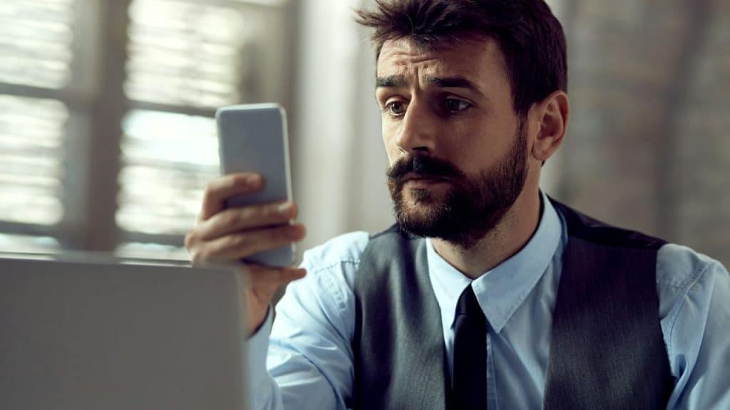 Man in a vest and tie looking intently at a smartphone in an office setting.